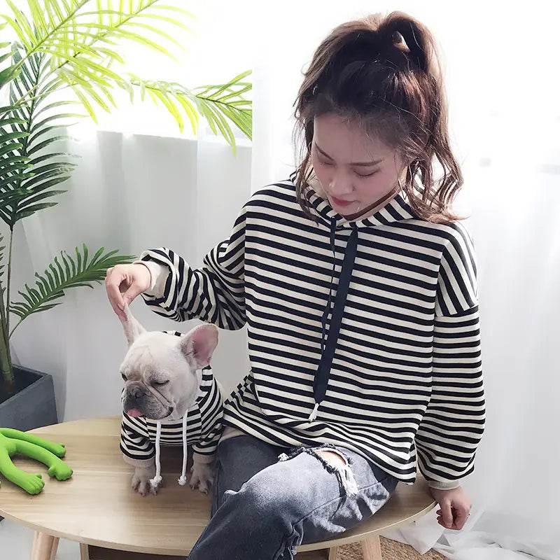 Woman in striped hoodie with French Bulldog wearing matching pet outfit, cozy indoor setting
